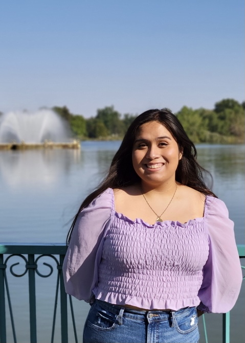 A headshot of me with a purple blouse trees and a lake.